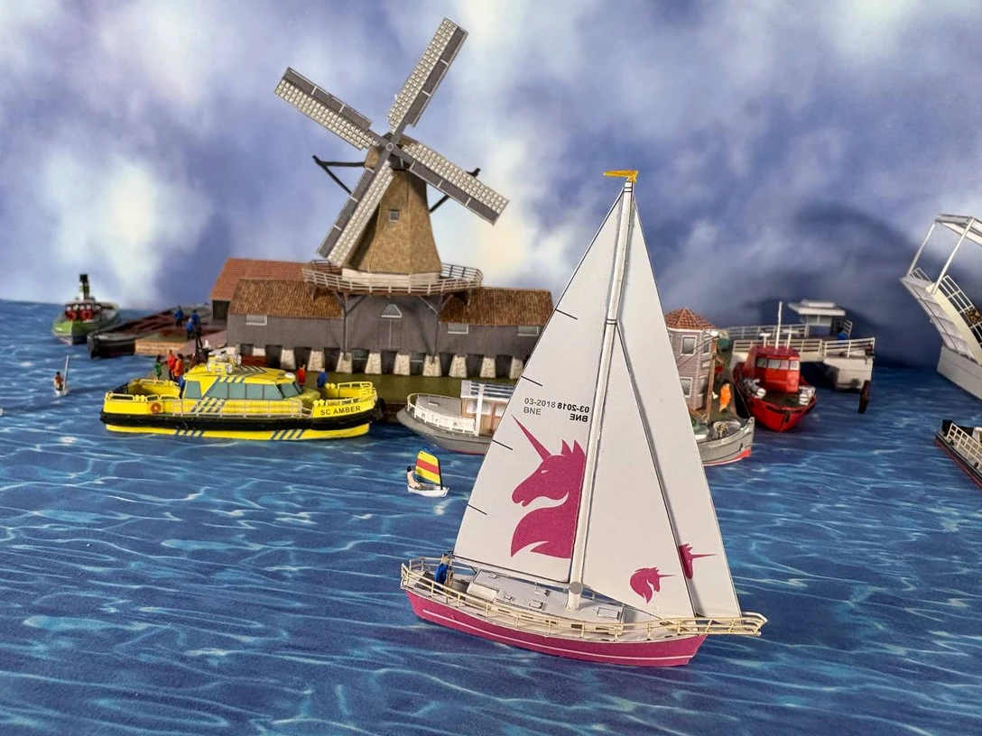 Karton-Modellbau Kartonmodell eines Segelbootes mit pinkem Einhornmotiv vor Mühle und weiteren Schiffen auf Wasserfläche.