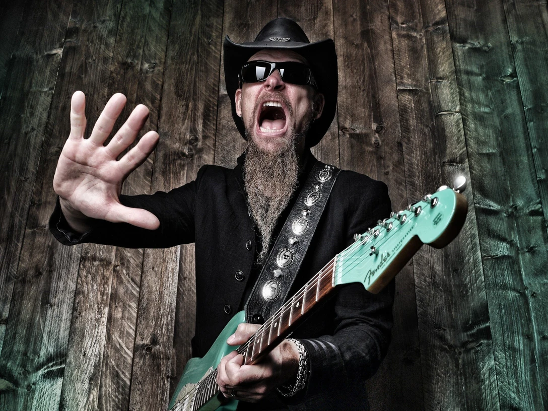 Dudley Taft Mann mit Bart, Hut und Sonnenbrille spielt E-Gitarre und streckt die Hand in Richtung Kamera.