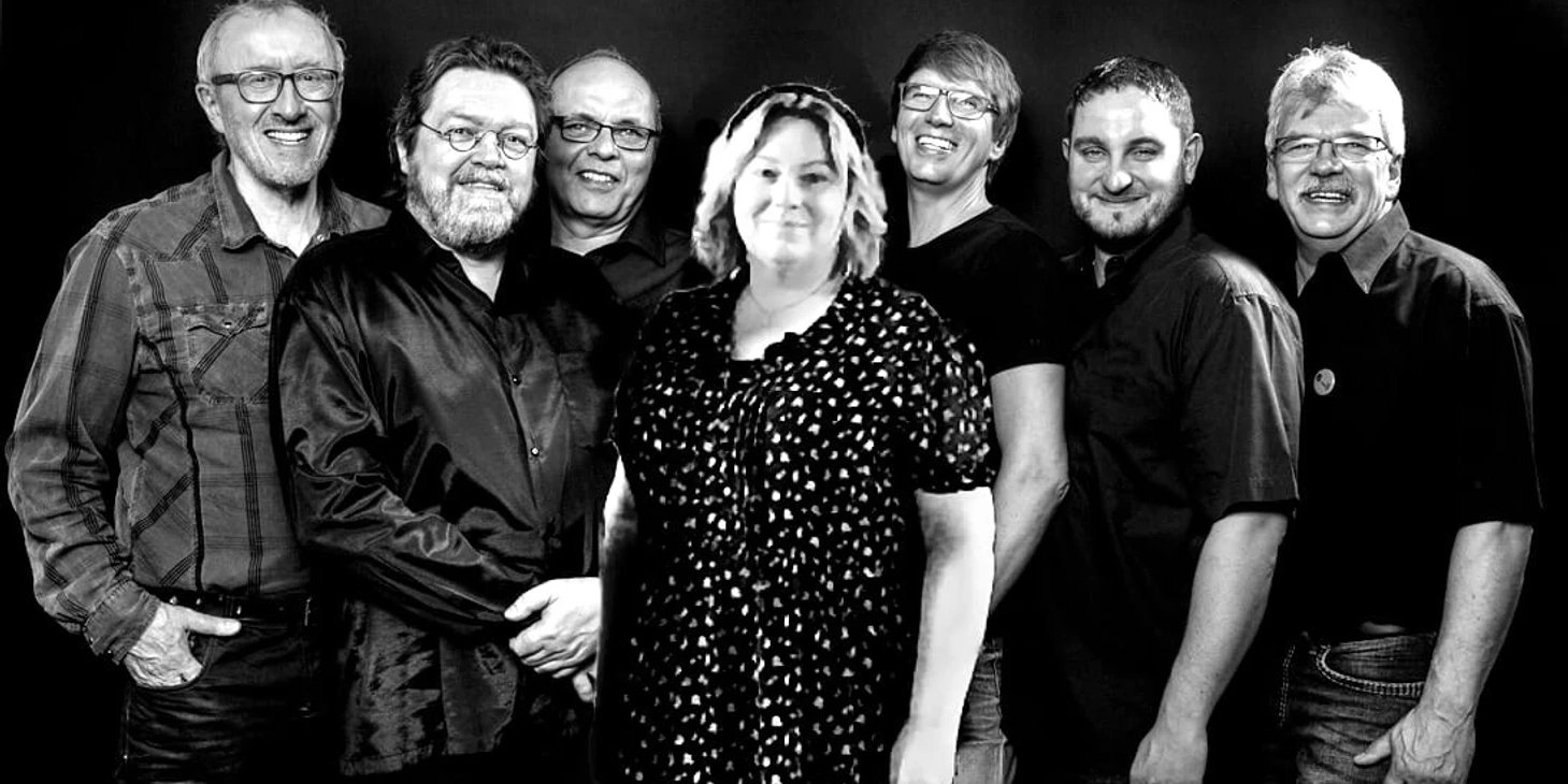 Six-and-One Acht Personen stehen eng beieinander und lächeln in die Kamera vor dunklem Hintergrund.