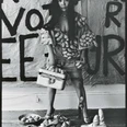 Yayoi Kusama posiert 1971 in ihrem New Yorker Atelier in Untitled (Dress) mit ihren phallischen Skulpturen Eine Frau posiert in einem Gewand mit knotigen Formen, umgeben von Schuhen und einem Koffer.