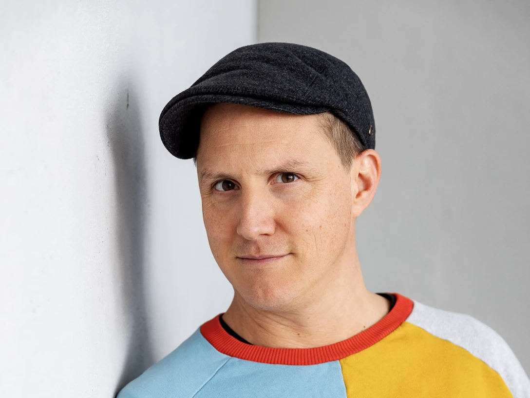 Sebastian-23 Person mit dunkler Schirmmütze und buntem Pullover blickt freundlich in die Kamera vor grauer Wand.