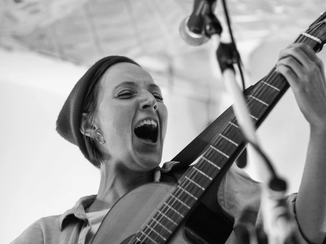 Lia L. Shoshann Musikerin mit Gitarre singt leidenschaftlich in ein Mikrofon auf einer Bühne.