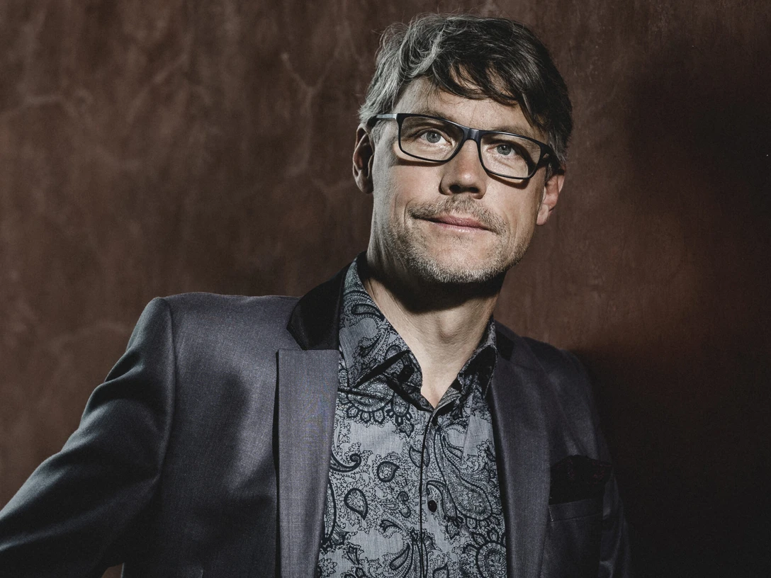 Christoph Sieber Mann mit Brille im Anzug vor braunem Hintergrund, blickt ruhig in die Kamera.