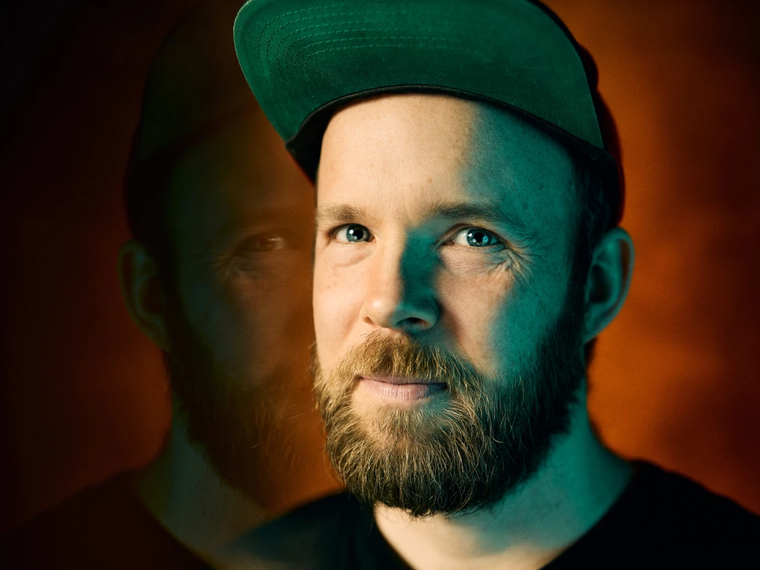 Quichotte – Alles echt Mann mit Bart und grüner Kappe blickt in die Kamera, sein Gesicht spiegelt sich neben ihm.