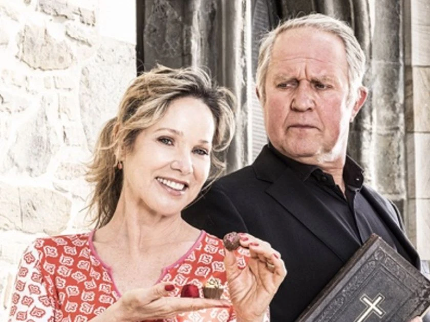 Chocolat - mit Harald Krassnitzer & Ann-Kathrin Kramer Frau mit Praline und lächelndem Gesicht steht neben einem Mann mit Buch vor steinener Wand.