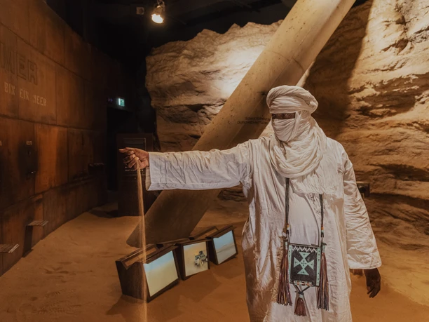 Klimahaus 4 Ibrahim Kane Annour-L (c) Hannes Voigts_Erlebnis Bremerhaven.jpg Mann in weißem Tuchgewand steht in sandiger Ausstellung und lässt Sand durch die Hand rieseln.