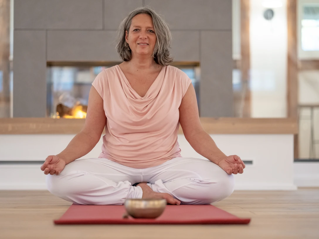 4 - DSC07588.jpg Frau sitzt entspannt im Schneidersitz auf einer Yogamatte vor einem Kamin und meditiert.