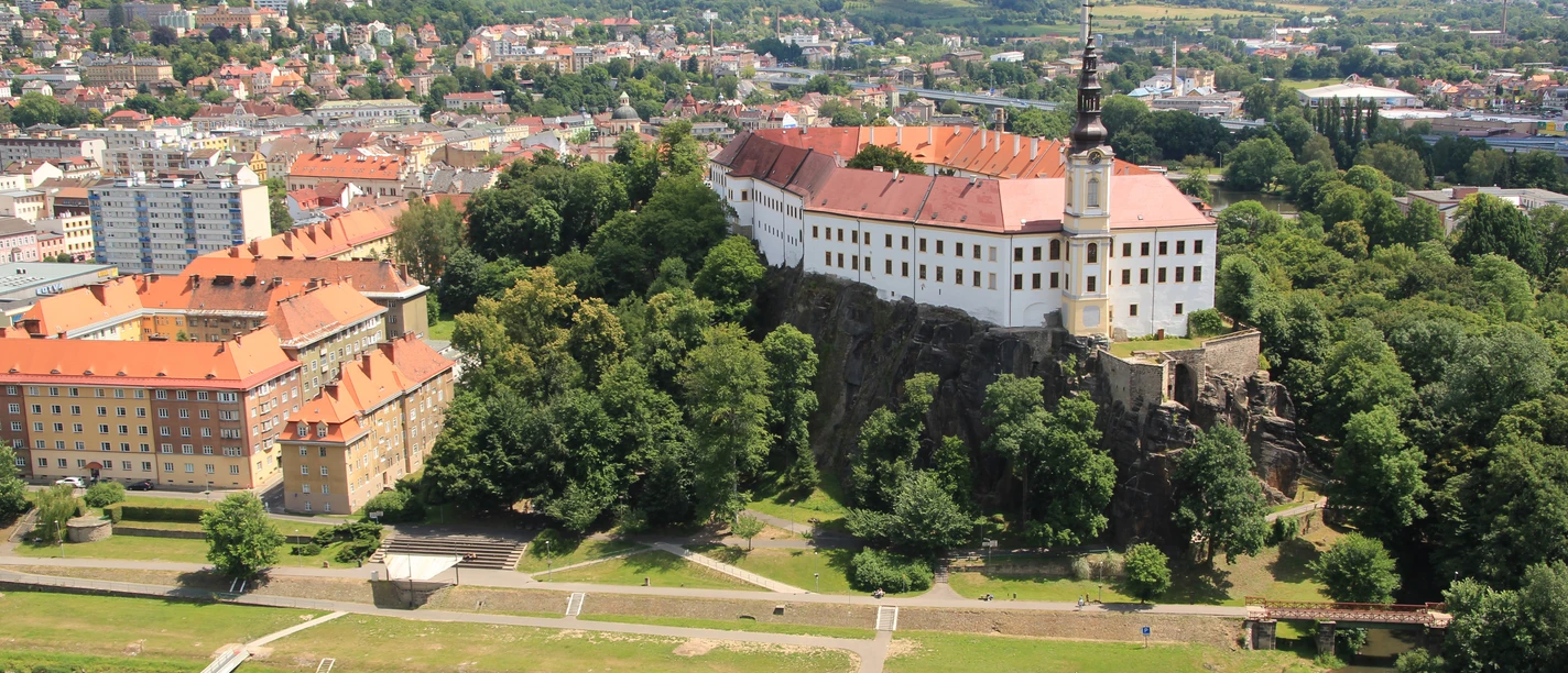 Schloss Děčín Luftaufnahme eines Schlosses auf einem Hügel neben einem Fluss mit Booten und Stadtansicht.