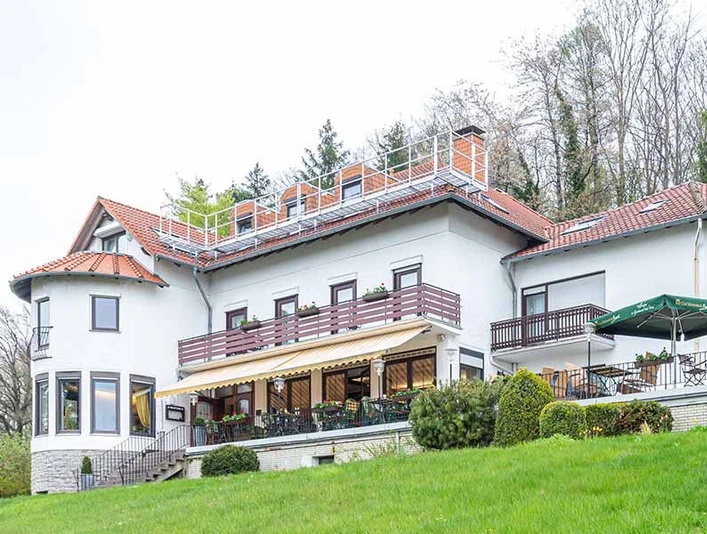 hotel_berghof_galerie_01.jpg Weißes Hotelgebäude mit rotem Dach, großer Terrasse, Sitzplätzen und Blick auf eine begrünte Anhöhe.