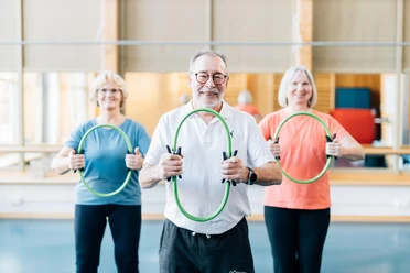 Drei ältere Erwachsene trainieren lächelnd mit grünen Fitnessringen im Kursraum