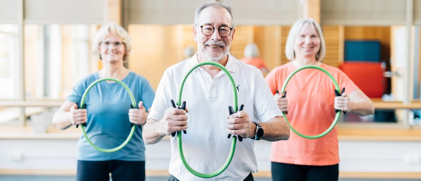 Drei ältere Erwachsene trainieren lächelnd mit grünen Fitnessringen im Kursraum