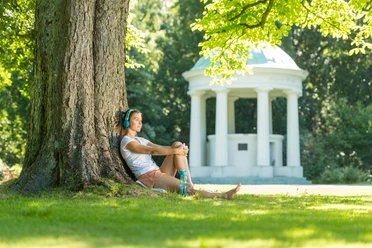 Eine junge Frau sitzt entspannt unter einem Baum und genießt die Natur. Im Hintergrund ist ein klassischer Pavillon zu sehen.