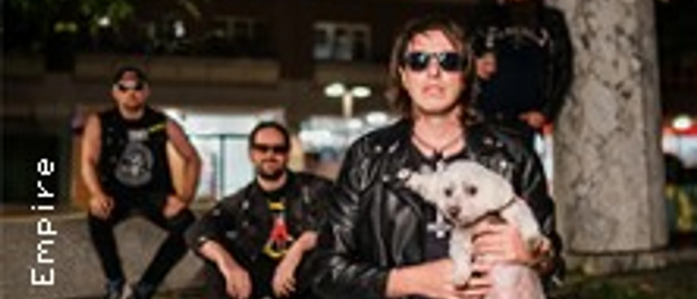 Vier Personen in dunkler Kleidung, eine Person hält einen kleinen Hund auf einem Betonmauerstück.