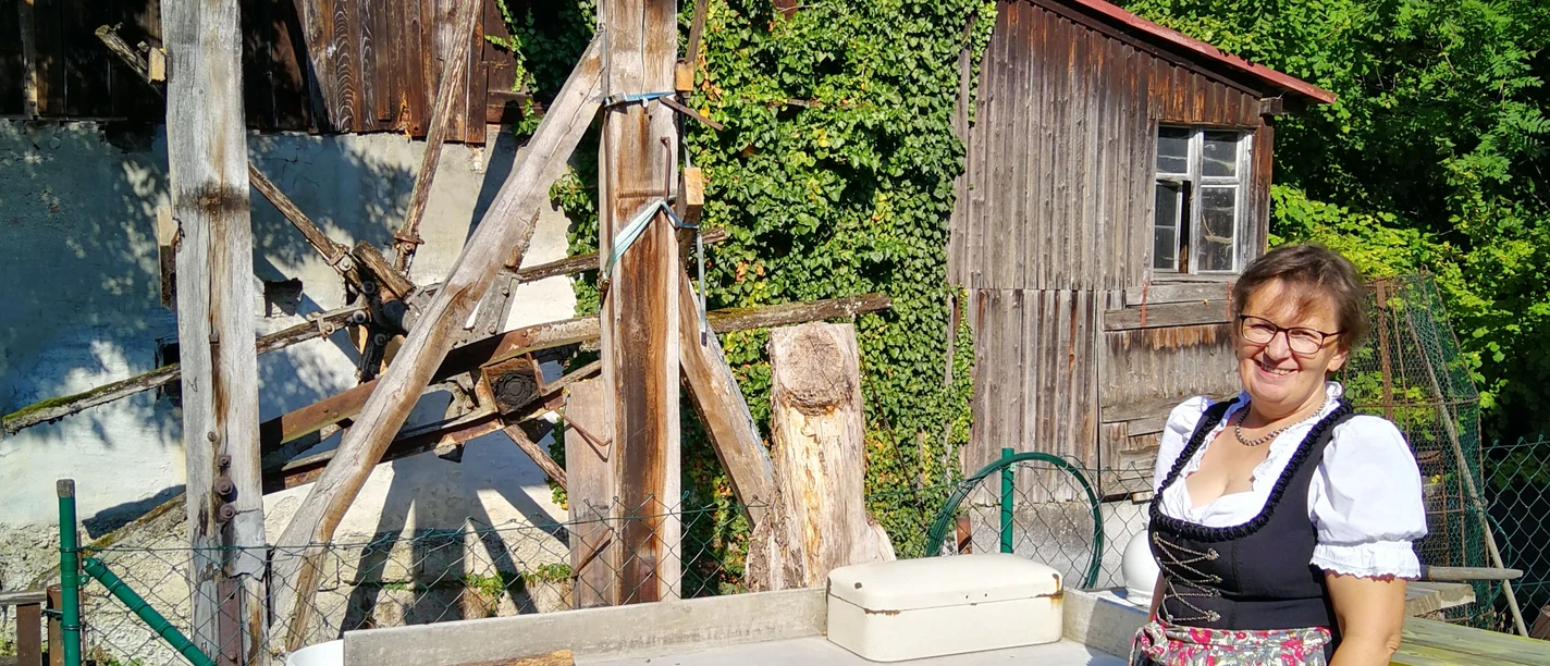 Wasserläufe, Mühlen und Industrie -Die Wasserführung Frau in traditioneller Tracht steht lächelnd vor alter Holz-Mühle mit grünen Pflanzen im Hintergrund.