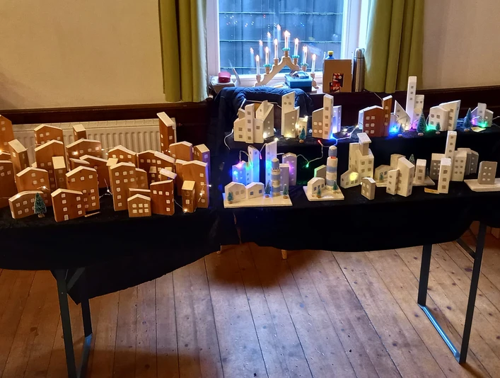 Tisch mit selbstgebastelten Geschenkideen Holzmodelle von Häusern und beleuchtete Stadtsilhouetten auf zwei Tischen in einem hellen Raum.