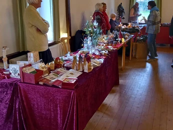 Adventsbasar im Schützenhof (2).jpg Menschen stehen an liebevoll geschmückten Tischen mit Kunsthandwerk und Geschenkartikeln in einer Halle.