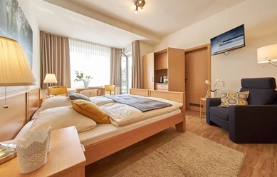 Haus am Kurpark Helles Doppelzimmer mit Holzmöbeln, Sessel, Fernseher, kleiner Küchenzeile und großem Fenster.
