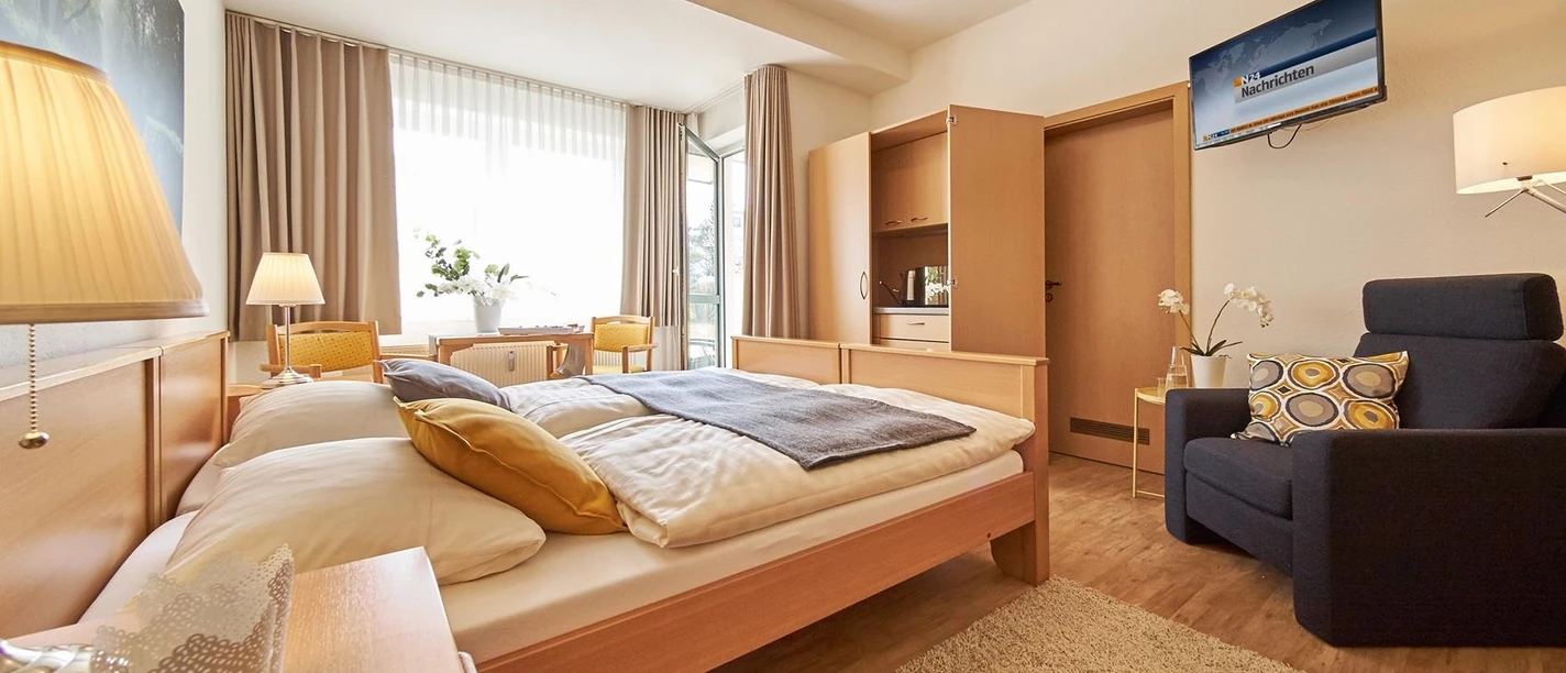 Haus am Kurpark Helles Doppelzimmer mit Holzmöbeln, Sessel, Fernseher, kleiner Küchenzeile und großem Fenster.