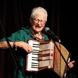 Ältere Frau spielt Akkordeon und singt in ein Mikrofon auf der Bühne.