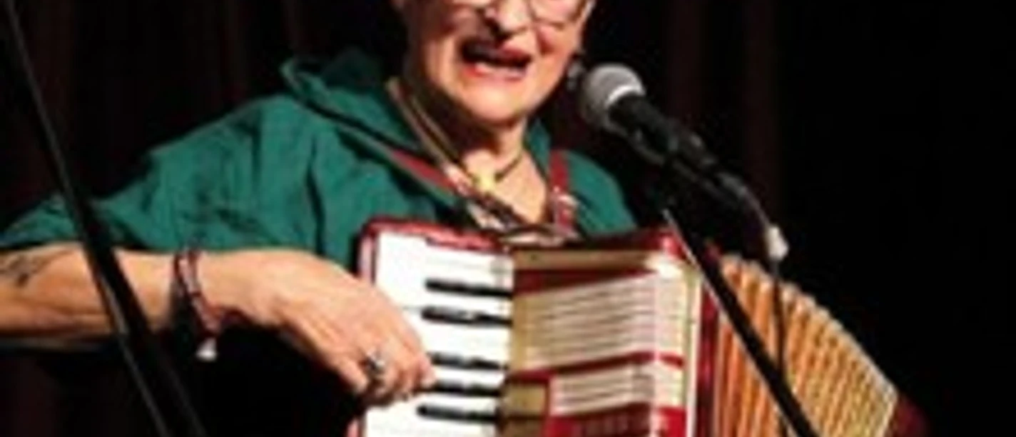 Ältere Frau spielt Akkordeon und singt in ein Mikrofon auf der Bühne.