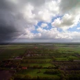 Schatteburg_BlickzumPolder_052019_kleiner.jpg Luftaufnahme einer grünen Landschaft mit Feldern und vereinzelten Gebäuden unter einem bewölkten Himmel mit dunklen und hellen Wolken.