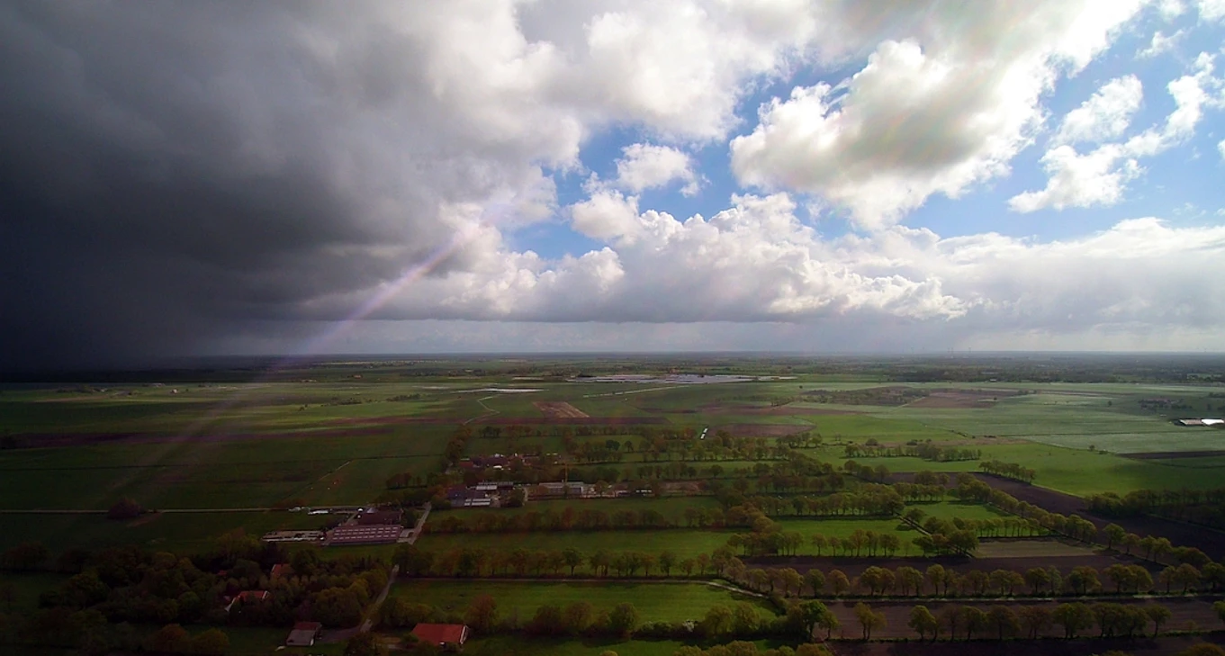 Schatteburg_BlickzumPolder_052019_kleiner.jpg Luftaufnahme einer grünen Landschaft mit Feldern und vereinzelten Gebäuden unter einem bewölkten Himmel mit dunklen und hellen Wolken.