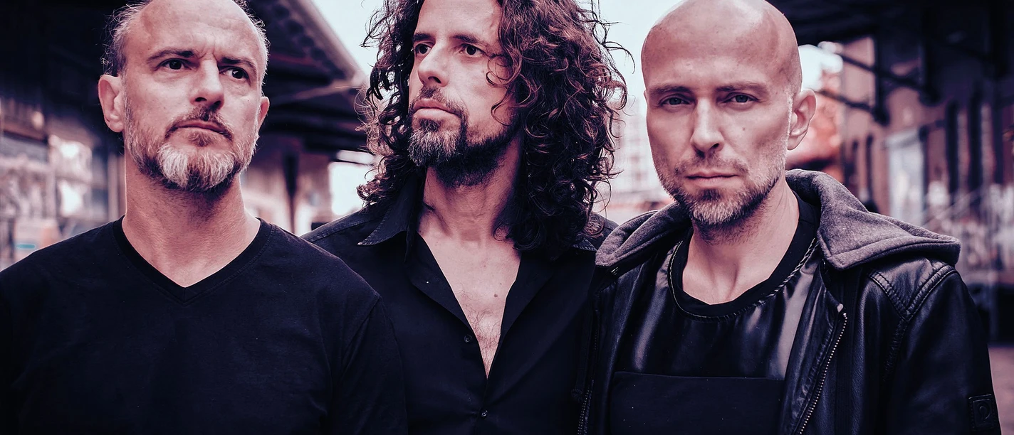 forced-to-mode-niko-schmuck-quer-cmyk-2.jpg Drei Männer in dunkler Kleidung stehen nebeneinander vor urbaner Kulisse mit Backsteinfassaden