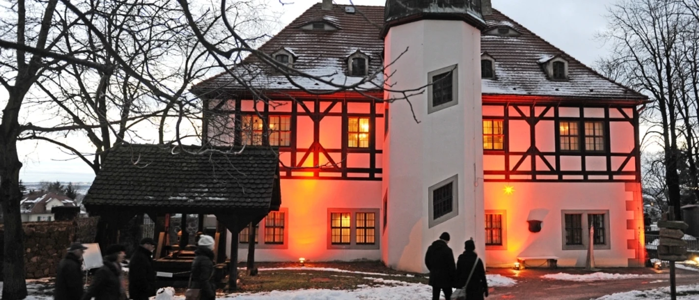 Hoflößnitzer_Weihnacht_Stiftung_Hofloessnitz.jpg Menschen folgen einem verschneiten Weg zu einem rot erleuchteten Fachwerkgebäude