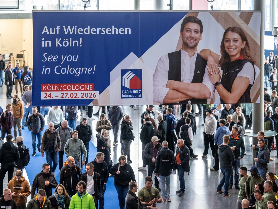 Geschäftiger Messebereich mit einem großen Banner einer Veranstaltung und zahlreichen Menschen.Busy trade fair area with a large event banner and lots of people.