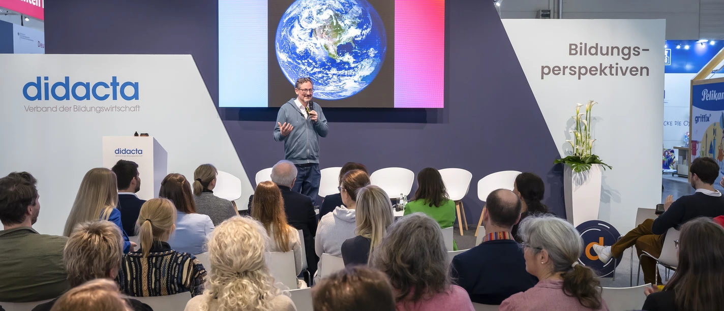 A speaker is standing in front of an audience in a conference room. The earth can be seen on the screen.