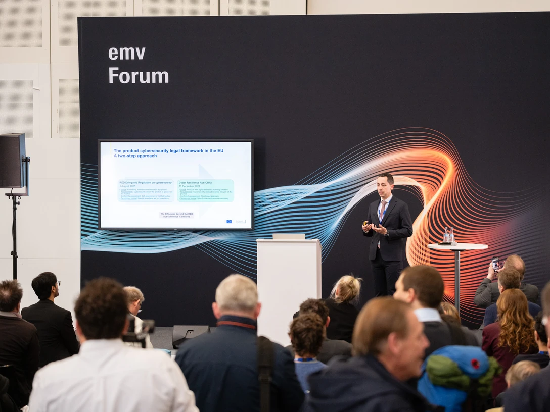 Ein Konferenzredner präsentiert einem Publikum Inhalte auf einer Messe mit dem Titel "emv Forum".A conference speaker presents content to an audience at a trade fair entitled "emv Forum".