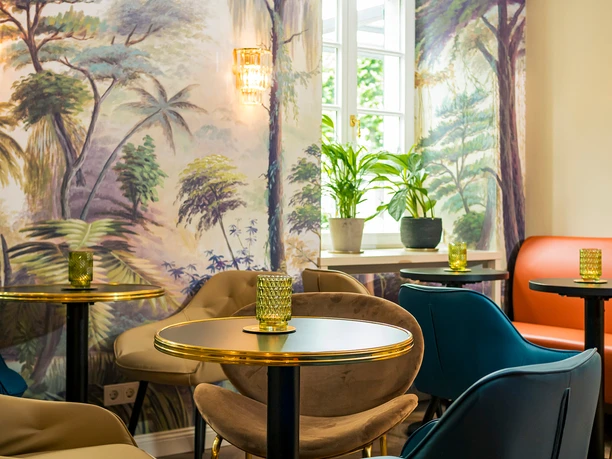 Altenberger Hof Hotel Sitzbereich mit runden Tischen, farbigen Sesseln, Pflanzen am Fenster und wandfüllendem Naturmotiv.