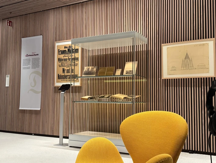 Blick in die Ausstellung "Wegbereiterinnen des Wandels" Eine Ausstellungsvitrine mit beleuchteten historischen Dokumenten und Bildern in einem modernen Raum.An exhibition showcase with illuminated historical documents and pictures in a modern room.