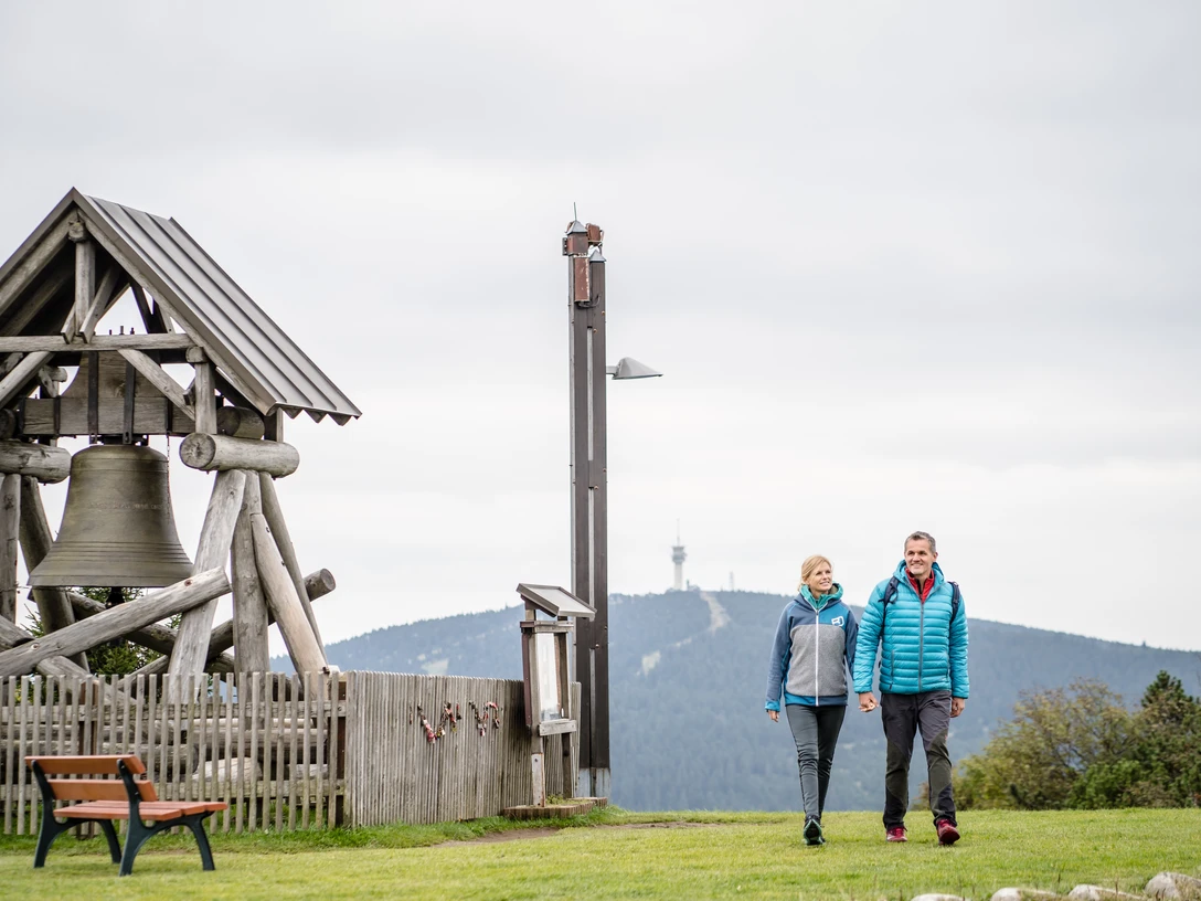 Erzgebirge_Oberwiesenthal_CC0_c_TMGS_Dennis_Stratmann (50).jpg Zwei Personen wandern neben einer großen Glocke in einer Berglandschaft.Two people are walking next to a large bell in a mountain landscape.Dva lidé jdou vedle velkého zvonu v horské krajině.Dwie osoby idą obok dużego dzwonu w górskim krajobrazie.Twee mensen lopen naast een grote klok in een berglandschap.Due persone camminano accanto a una grande campana in un paesaggio di montagna.