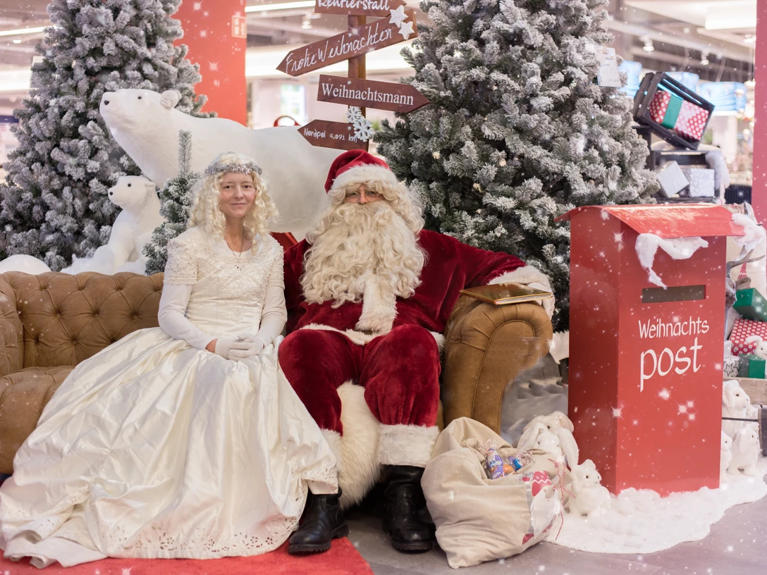 Weihnachstmann.jpg Weihnachtsmann und Engel sitzen vor geschmückten Tannenbäumen neben rotem Briefkasten für Wunschzettel.