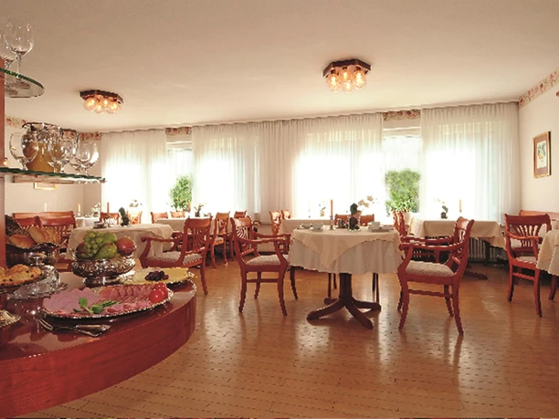 Speiseraum Heller Speiseraum mit Holzboden, gedeckten Tischen, Holzstühlen und Buffet mit Obst und Aufschnitt.