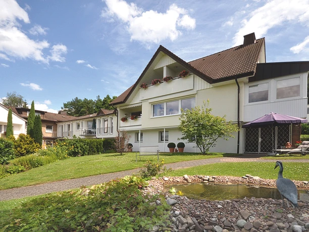 Außenansicht - Haus am Steinberg Helles Wohnhaus mit braunem Satteldach, gepflegtem Garten, Teich und Sitzbereich mit Sonnenschirm.