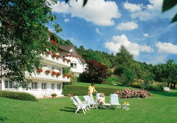 Haus am Steinberg Zwei Personen auf Liegestühlen vor einem weißen Haus mit Blumenbalkonen in grüner Gartenlandschaft.