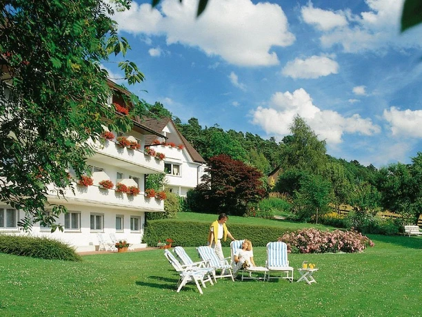 Haus am Steinberg Zwei Personen auf Liegestühlen vor einem weißen Haus mit Blumenbalkonen in grüner Gartenlandschaft.
