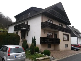Ferienwohnung Am Waldrand Außenansicht Ferienwohnung mit heller Fassade, dunklen Holzbalkonen und kleinem Vorgarten an der Straße.
