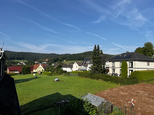 Ferienwohnung Mediterran Wohnhäuser am grünen Hang mit Wiese, Gewächshaus und Blick auf bewaldete Hügel unter blauem Himmel.