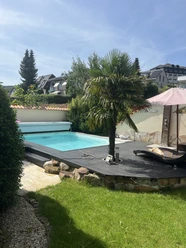 Ferienwohnung Mediterran Garten mit Pool, Holzterrasse, Palme und Liegestuhl unter Sonnenschirm bei sonnigem Wetter.