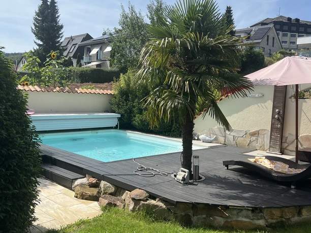 Ferienwohnung Mediterran Garten mit Pool, Holzterrasse, Palme und Liegestuhl unter Sonnenschirm bei sonnigem Wetter.