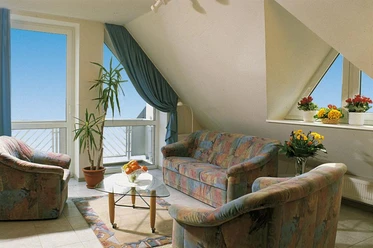 Gästehaus Therme-Blick Helles Wohnzimmer mit bunten Polstermöbeln, Glastisch, großen Fenstern und Blumen auf der Fensterbank.