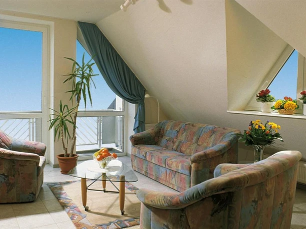 Gästehaus Therme-Blick Helles Wohnzimmer mit bunten Polstermöbeln, Glastisch, großen Fenstern und Blumen auf der Fensterbank.