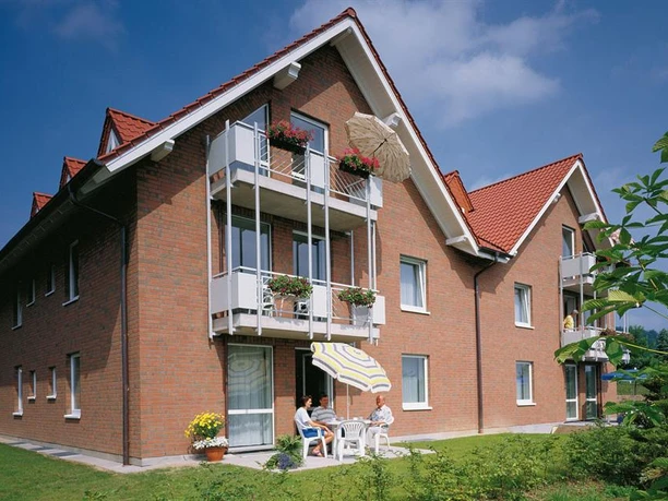 Gästehaus Therme-Blick Zweistöckiges Backsteinhaus mit roten Ziegeldächern, Balkonen, Terrasse und drei Personen im Garten.