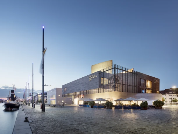 Deutsches Auswandererhaus Ensemble Hafenansicht 1 (c) Werner Huthmacher_Deutsches Auswandererhaus.jpg Moderne Museumsarchitektur am beleuchteten Hafenbecken mit Schiffen im Abendlicht von Bremerhaven.