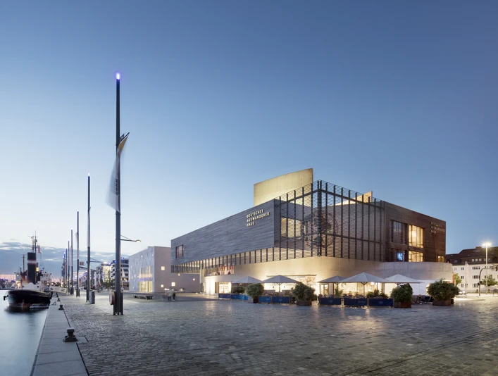 Deutsches Auswandererhaus Ensemble Hafenansicht 1 (c) Werner Huthmacher_Deutsches Auswandererhaus.jpg Moderne Museumsarchitektur am beleuchteten Hafenbecken mit Schiffen im Abendlicht von Bremerhaven.