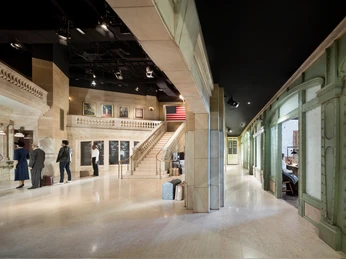 Deutsches Auswandererhaus Grand Central Terminal 6 (c) Werner Huthmacher_Deutsches Auswandererhaus.jpg Weitläufige Ausstellungshalle mit Marmortreppen, Reisenden, Gepäckstücken und US-Flagge an der Wand.