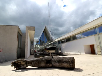 Deutsches Schifffahrtsmuseum IMG_4919 (c) Tanja Mehl_Erlebnis Bremerhaven.jpg Moderner Museumsbau mit Glasfassade, Segelboot und großer Holzskulptur im Außenbereich.
